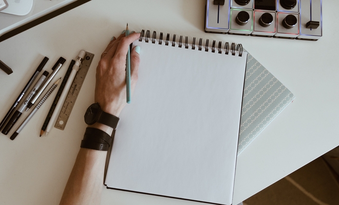 Image of a desk with a hand holding a pencil atop a blank notepad